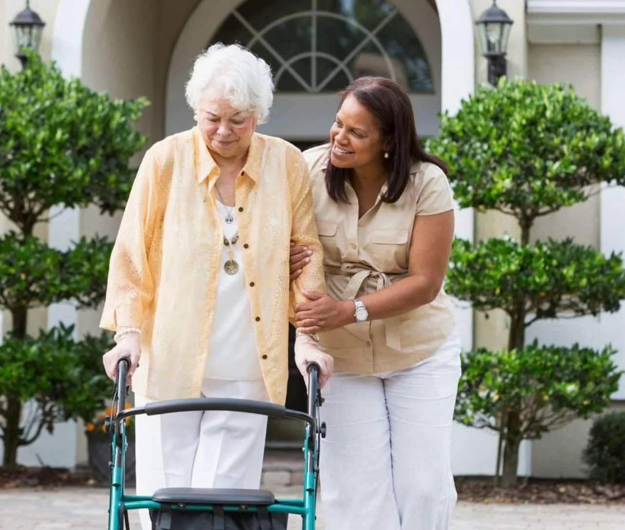 A young woman helping an elderly woman walk with a walker outside a house surrounded by green bushes.