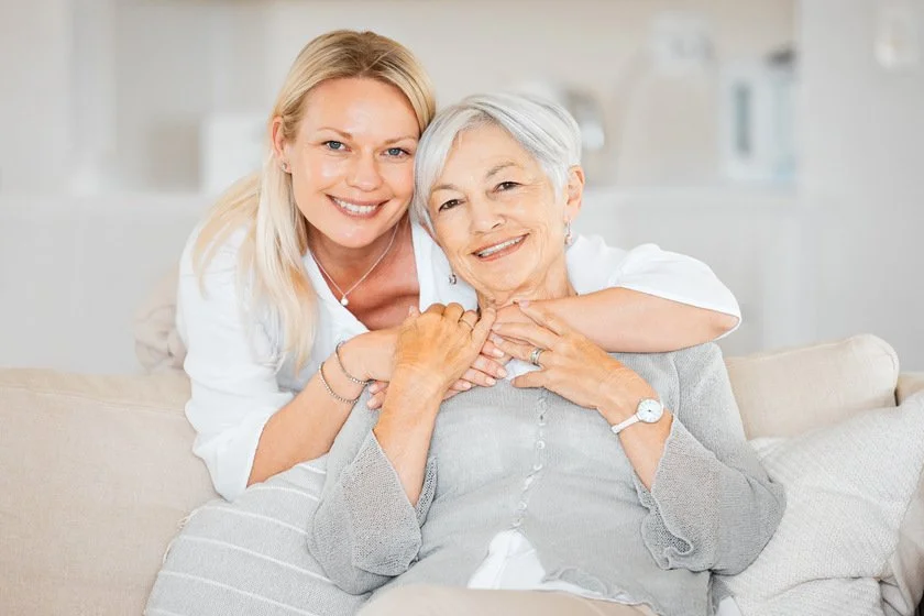 Two women, an elderly woman with gray hair and a younger woman with blonde hair, sitting and smiling together on a sofa in a bright living room.