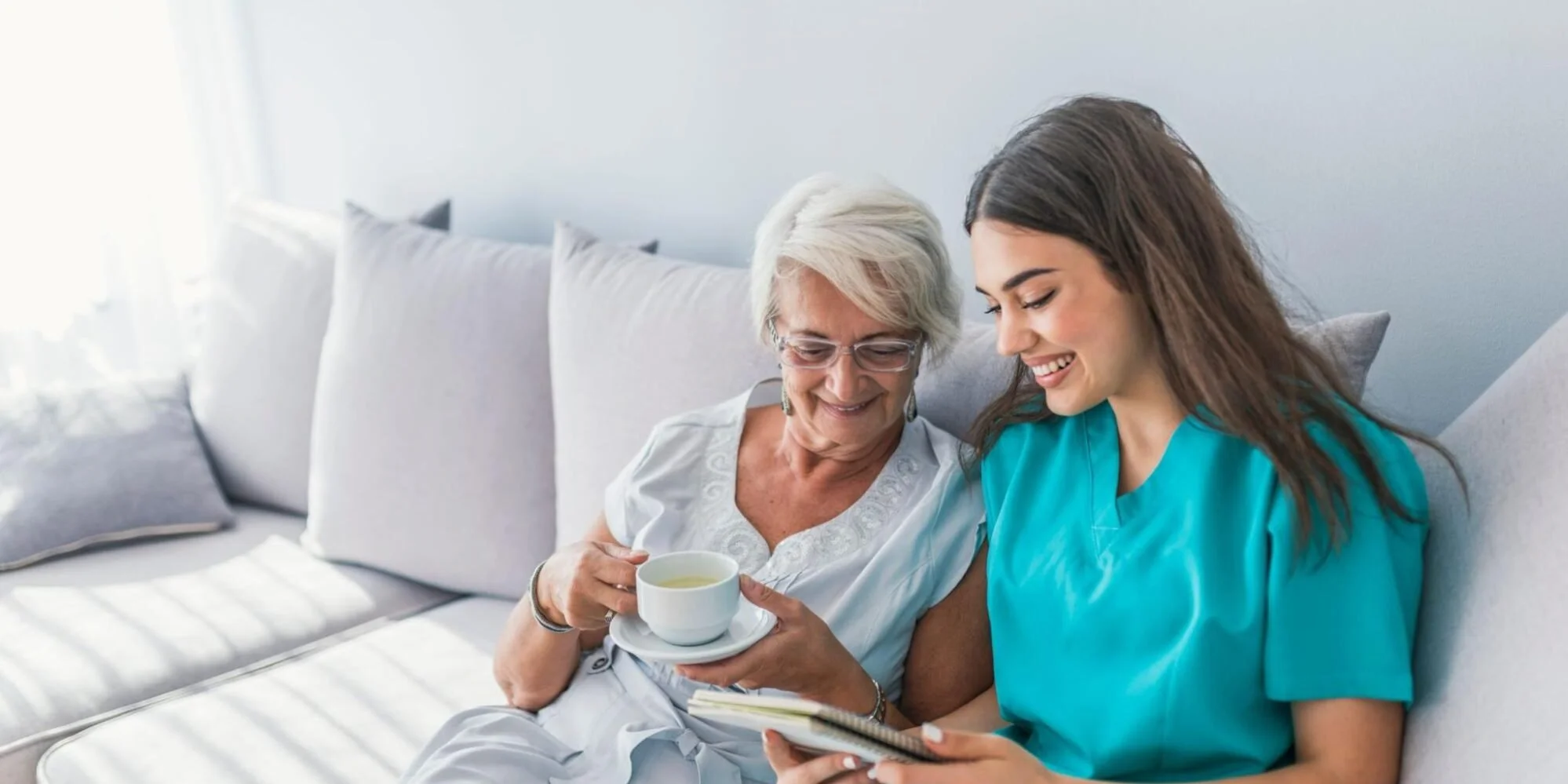 An elderly woman and a young woman in medical scrubs smiling on a beige sofa, the elderly woman holding a teacup, with sunlight coming through a window in the background.
