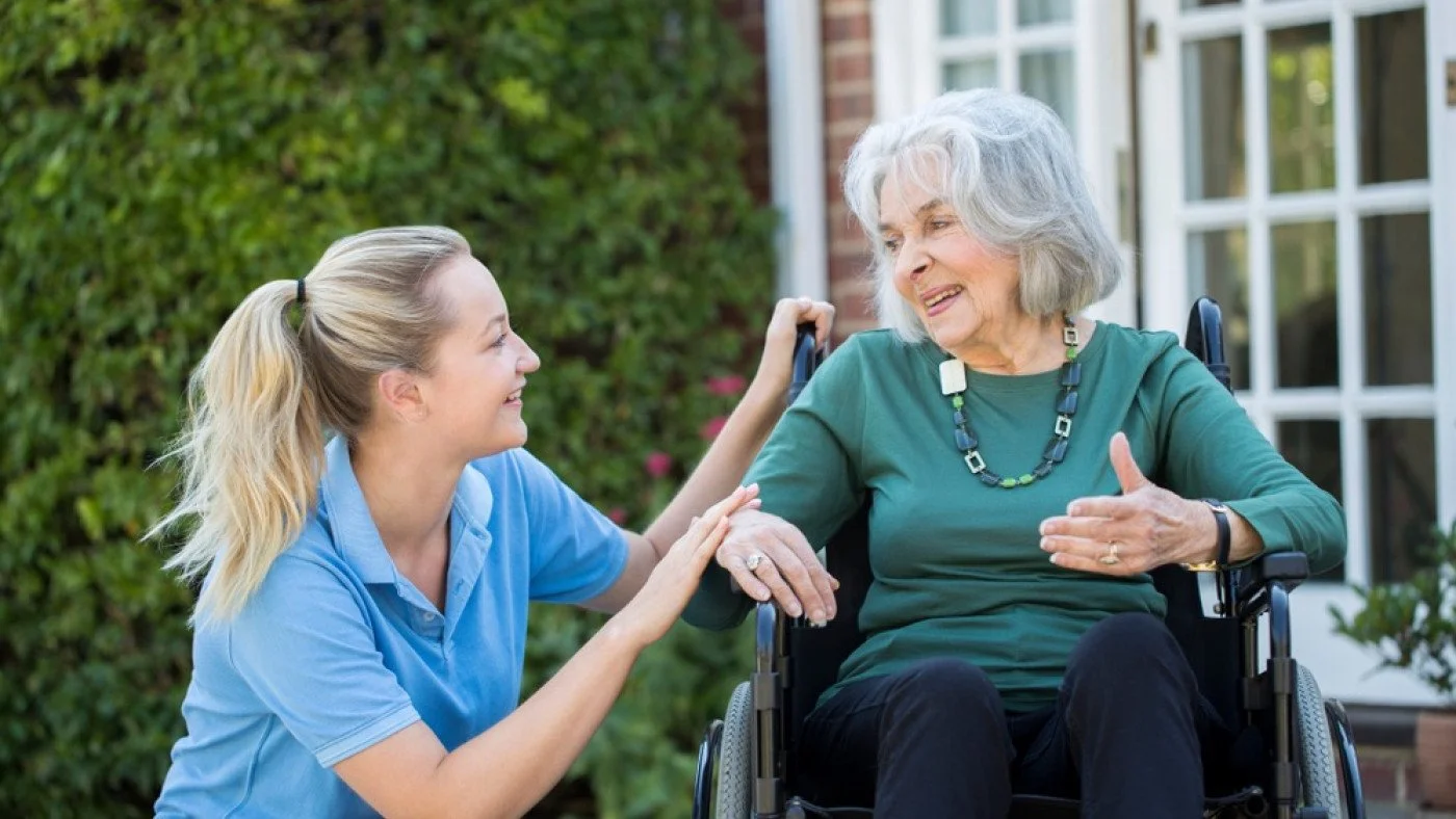 A young woman with blonde hair in a ponytail and a blue uniform is smiling and holding hands with an elderly woman with white hair in a wheelchair, outside a house with green bushes in the background.