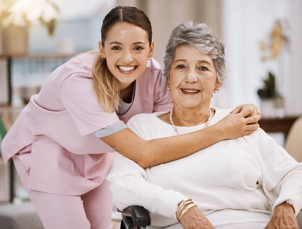 A young woman in pink scrubs smiling with an older woman in a wheelchair, both showing happiness.