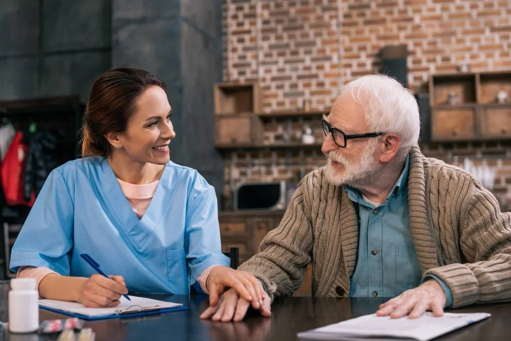 A young female nurse smiling and holding a pen, talking to an elderly man with glasses, wearing a brown cardigan, at a table in a room with brick walls and shelves. She appears to be taking notes or explaining something.