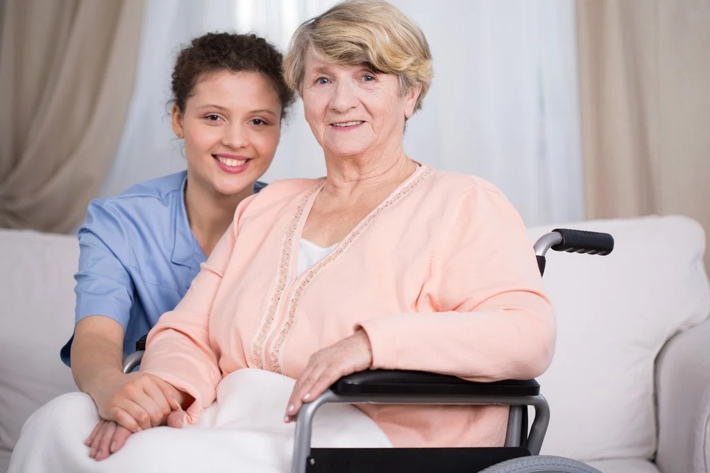A young woman in blue scrubs sitting next to an elderly woman in a wheelchair, both smiling indoors with curtains and a bed in the background.