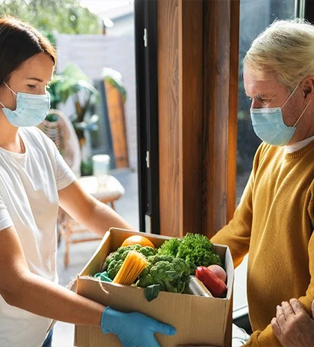 A woman wearing a face mask and gloves hands a box of fresh vegetables to an elderly woman wearing a face mask inside a house.