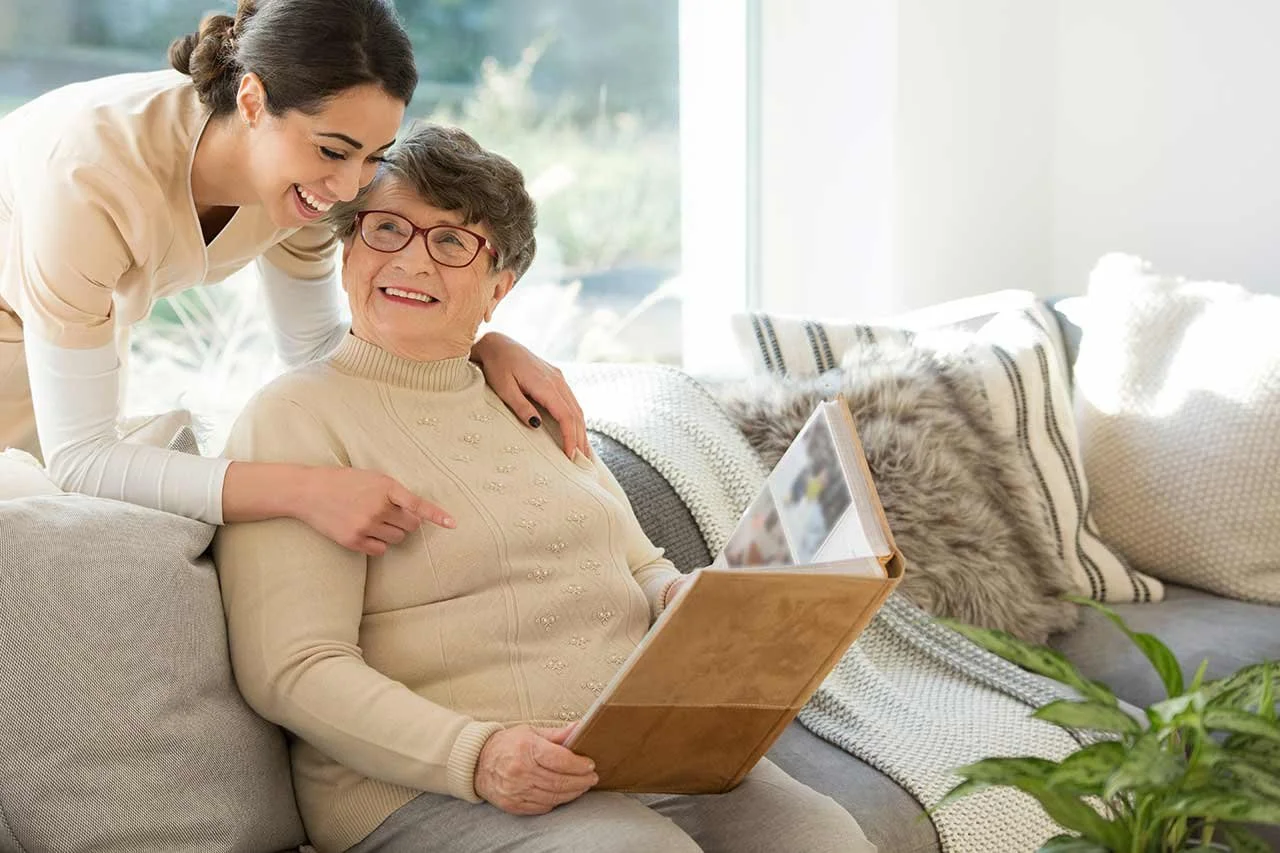 A young woman and an elderly woman sitting on a sofa, smiling and looking at a photo album together in a bright living room.