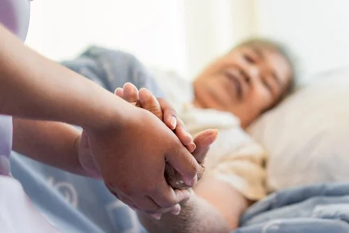 A person holding an elderly woman's hand in a hospital room.
