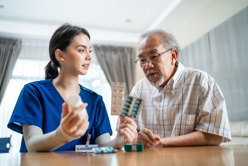 A young female nurse holding pills and a blister pack, talking to an elderly man with glasses who is also holding medications, in a brightly lit room.