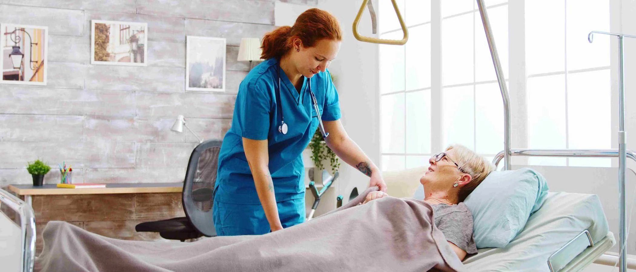 Nurse caring for an elderly woman patient in a hospital bed, holding her hand and smiling, in a bright room with large windows and wall decorations.