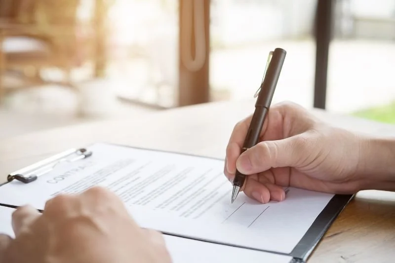 Person signing a contract on a clipboard with a pen, with a blurred background of a window and natural light.
