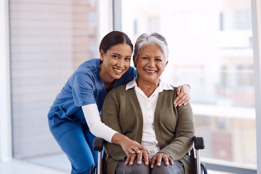 A healthcare worker in blue scrubs and a senior woman in a wheelchair smiling together in a bright room.