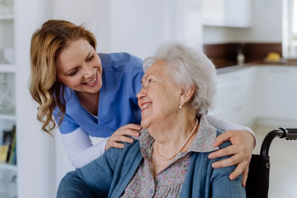 A young female healthcare worker smiling and comforting an elderly woman with white hair, wearing a plaid shirt, pearl necklace, and blue cardigan, who is sitting in a wheelchair in a bright room.