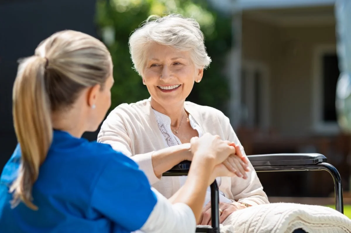 A young woman in blue scrubs holding the hands of an elderly woman with gray hair, smiling outdoors in a sunny setting.
