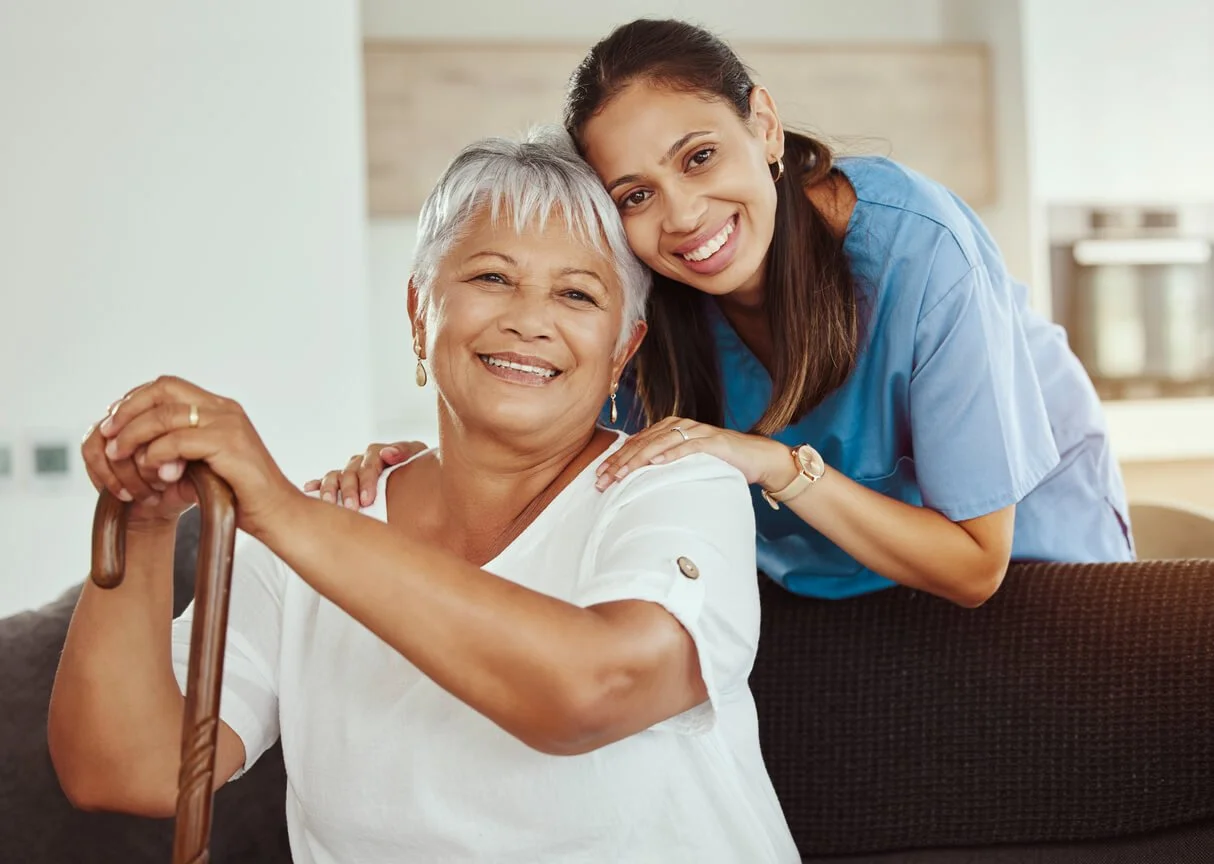 A smiling senior woman with short gray hair sitting with a cane, and a young female healthcare worker in blue scrubs standing behind her, both smiling and looking at the camera in a bright room.