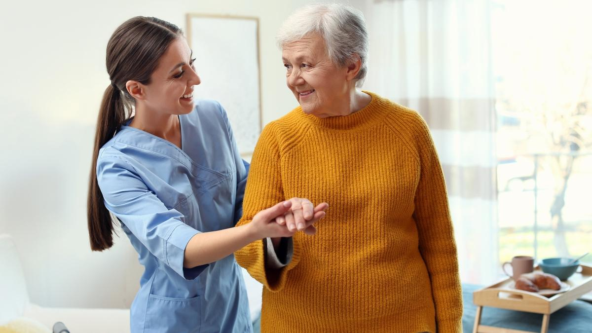A young female caregiver in light blue scrubs helping an elderly woman in a mustard yellow sweater walk inside a bright, sunny room.
