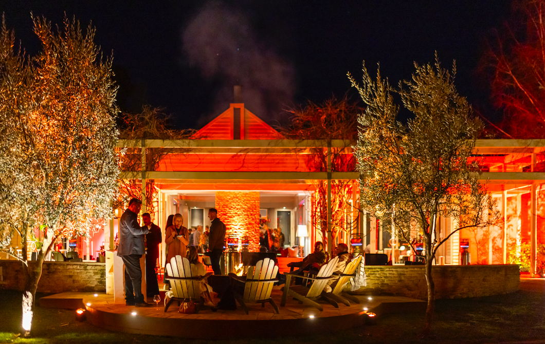 Nighttime outdoor gathering with people socializing on a patio, decorated with trees and warm lighting, in front of a lit house with a chimney and smoke