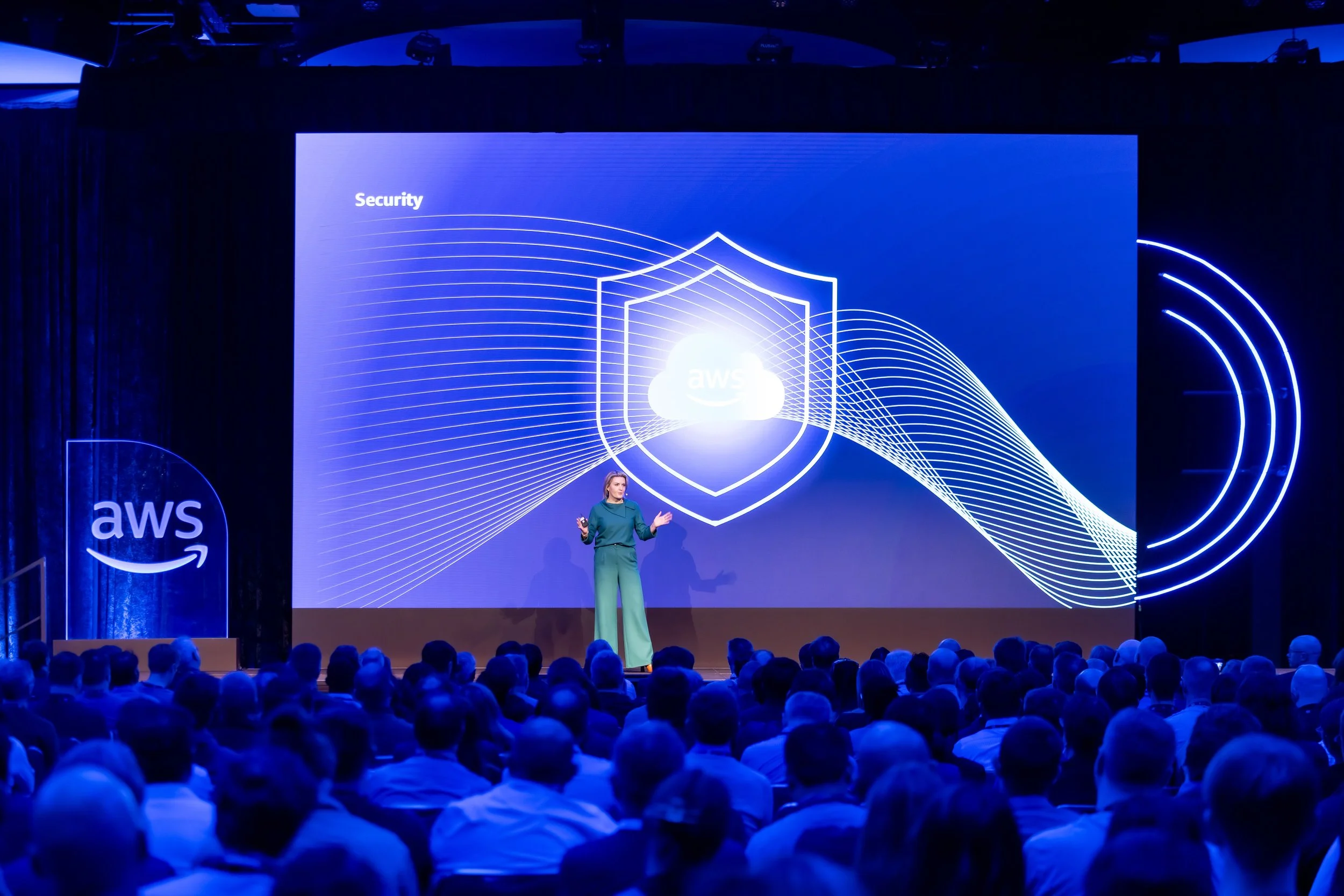A woman presenting on stage at an AWS conference with a large screen behind her displaying a security shield graphic with the AWS logo in the center and the word 'Security' in the top left corner. The audience is seated in front.