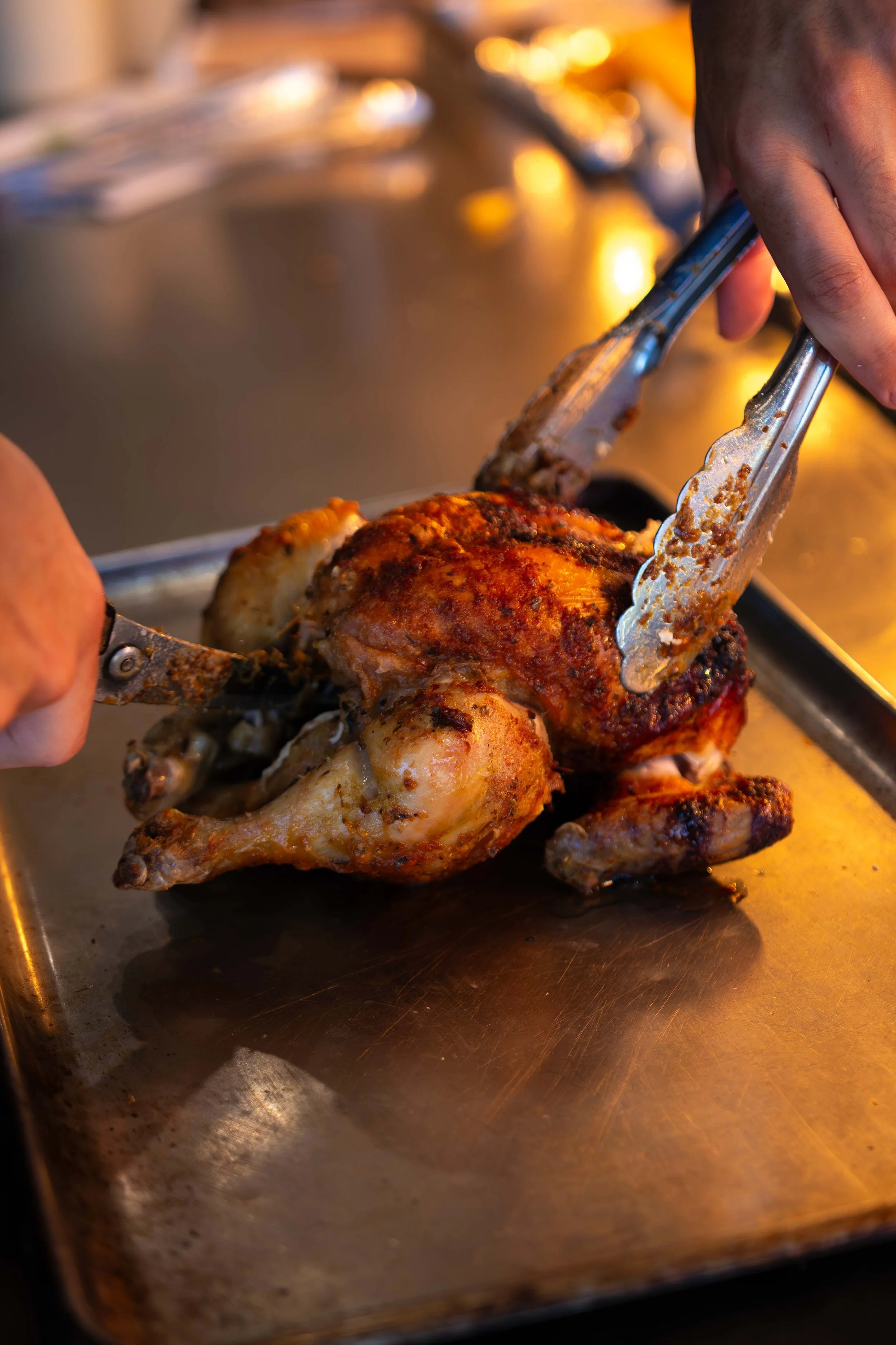 A person carving a roasted chicken with tongs on a metal tray.