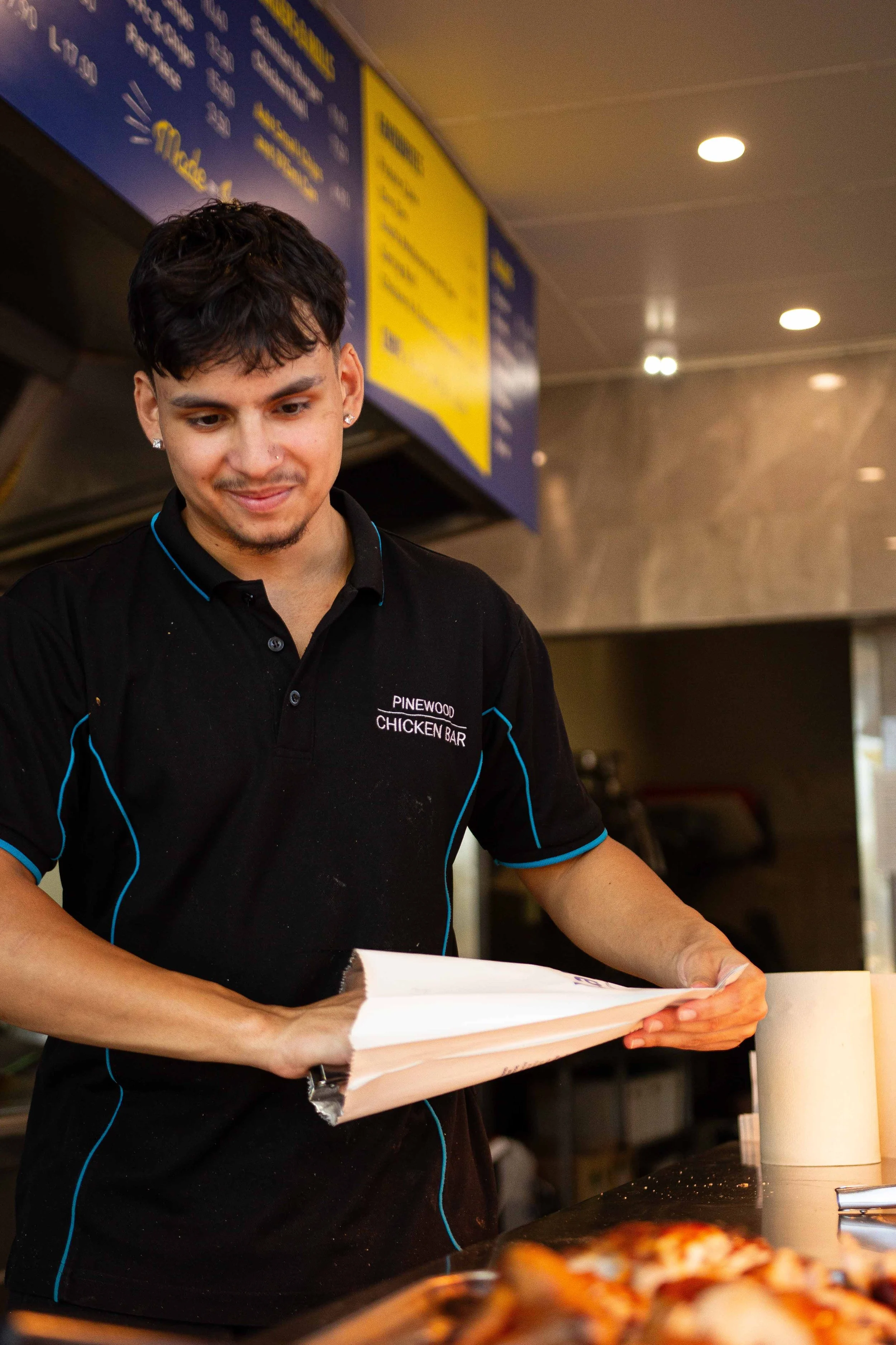 A young man in a black Pinewood Chicken Bar uniform is preparing a pizza in a restaurant.