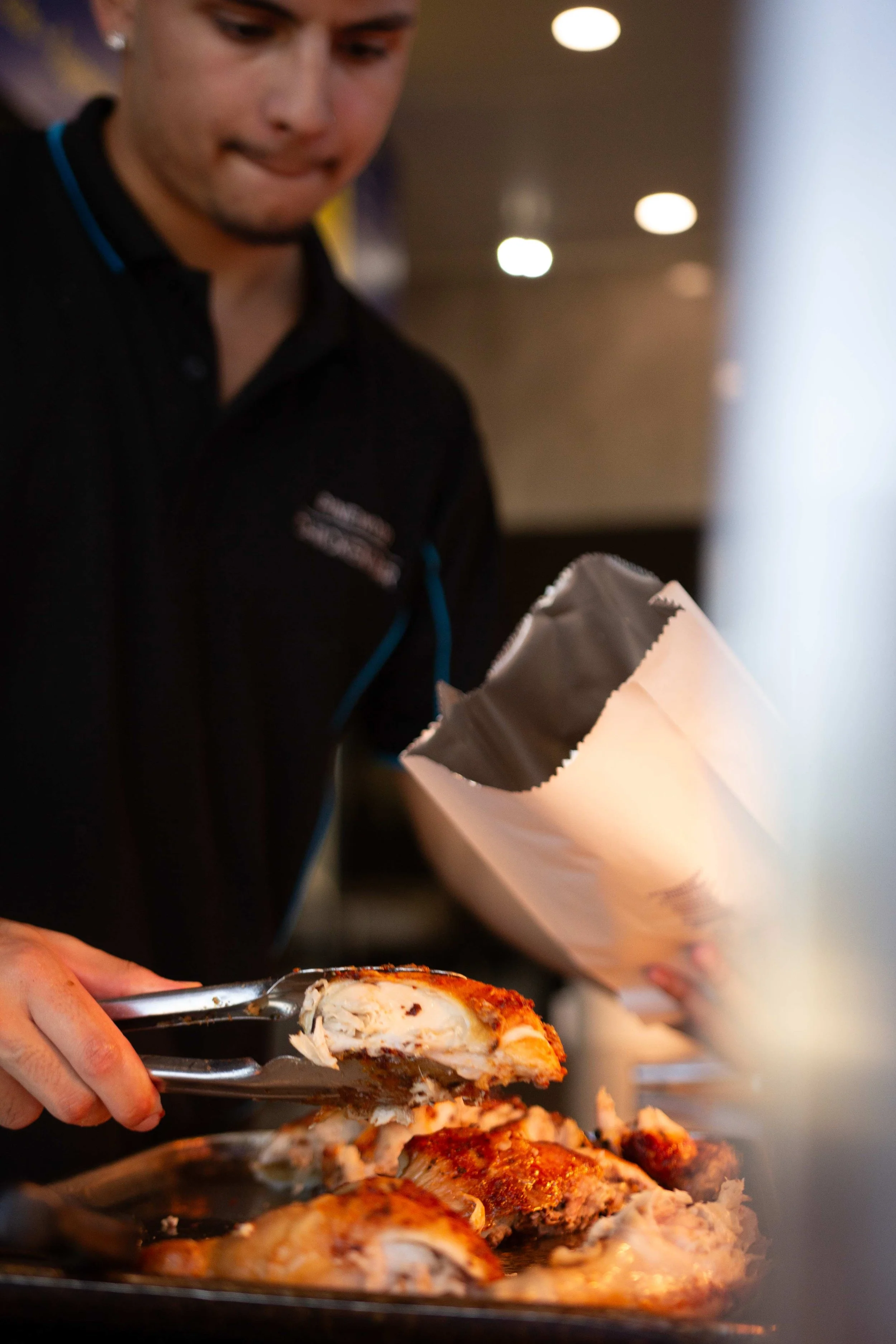 A person wearing a black shirt is serving slices of roasted chicken onto a plate, with the focus on the chicken and tongs.