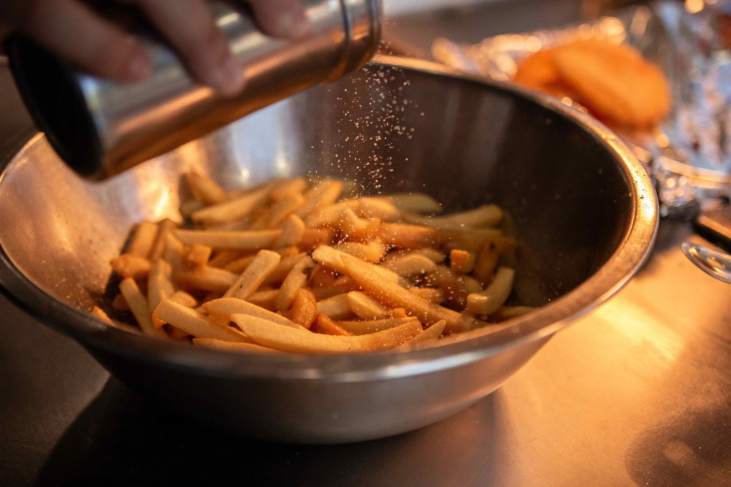 French fries being seasoned with salt in a metal bowl.