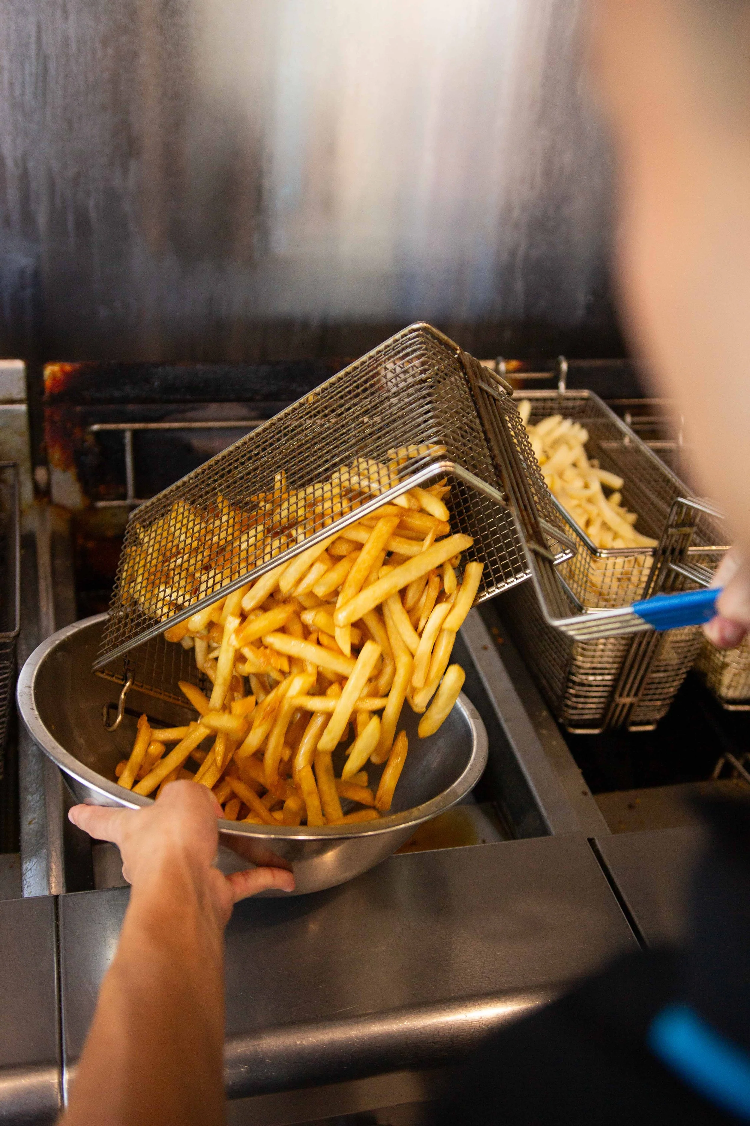 Someone is pouring cooked French fries from a metal basket into a metal bowl in a restaurant kitchen.