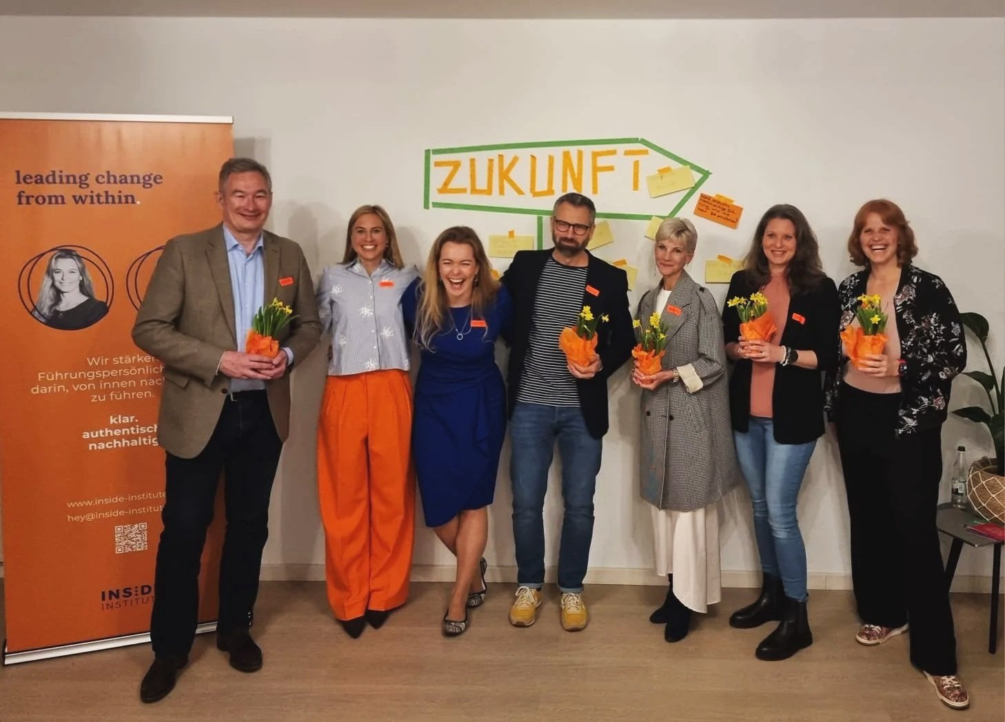 Gruppenzusammenstellung von sieben Menschen, die bei einer Veranstaltung stehen, einige halten Blumentöpfe mit gelben Blumen. Hinter ihnen ist ein Wand mit der Aufschrift 'ZUKUNFT' und Post-its. Rechts befindet sich ein orangefarbenes Banner mit Text und einem QR-Code.