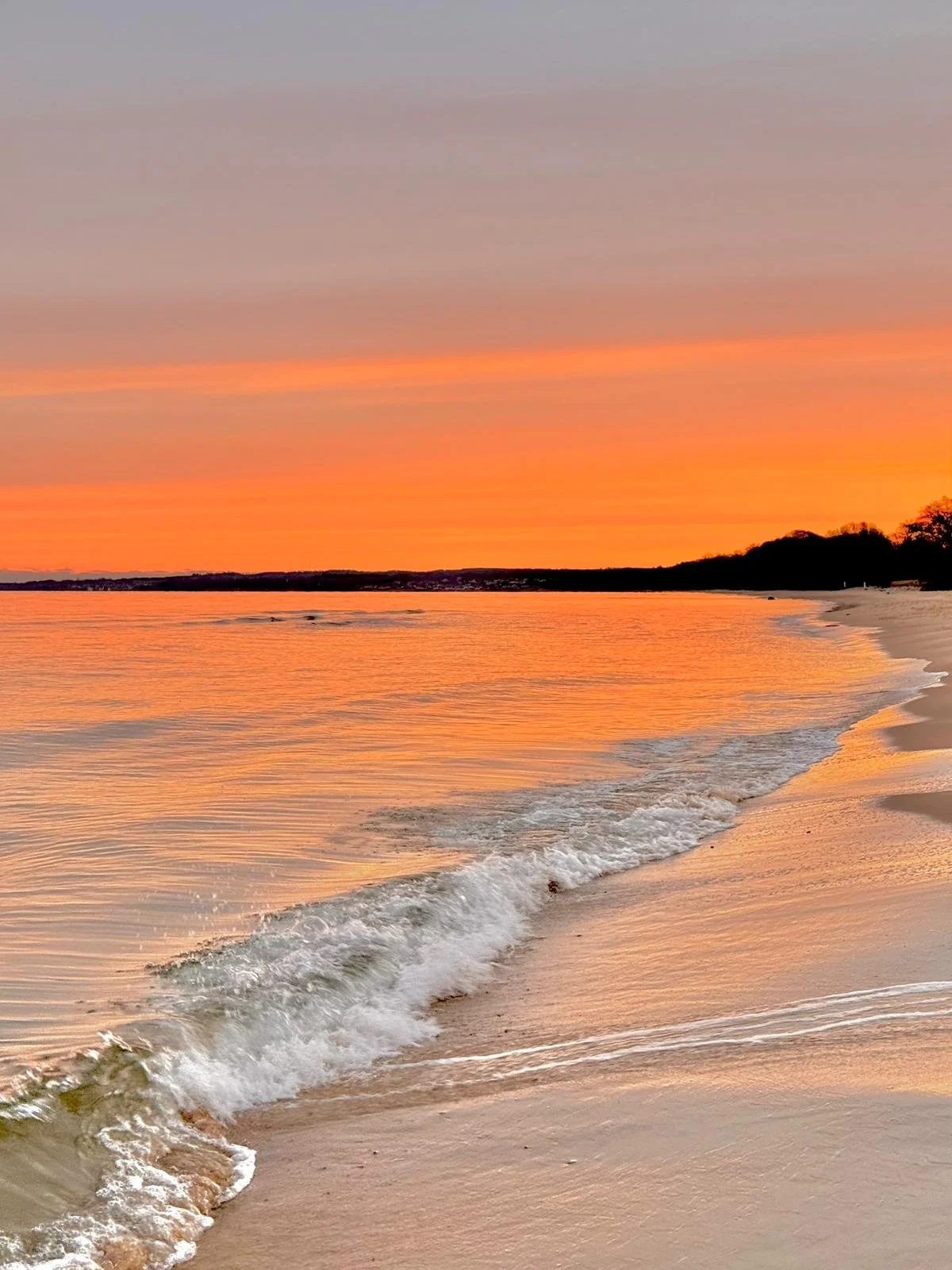 Ein Strand bei Sonnenuntergang mit sanften Wellen, sandigem Ufer und einem Himmel in Orangetönen.