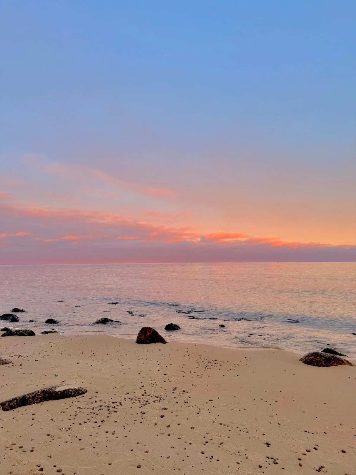 Ein ruhiger Strand bei Sonnenuntergang mit Sand, Steinen und dem Meer im Hintergrund. Der Himmel ist in sanften Pastellfarben.