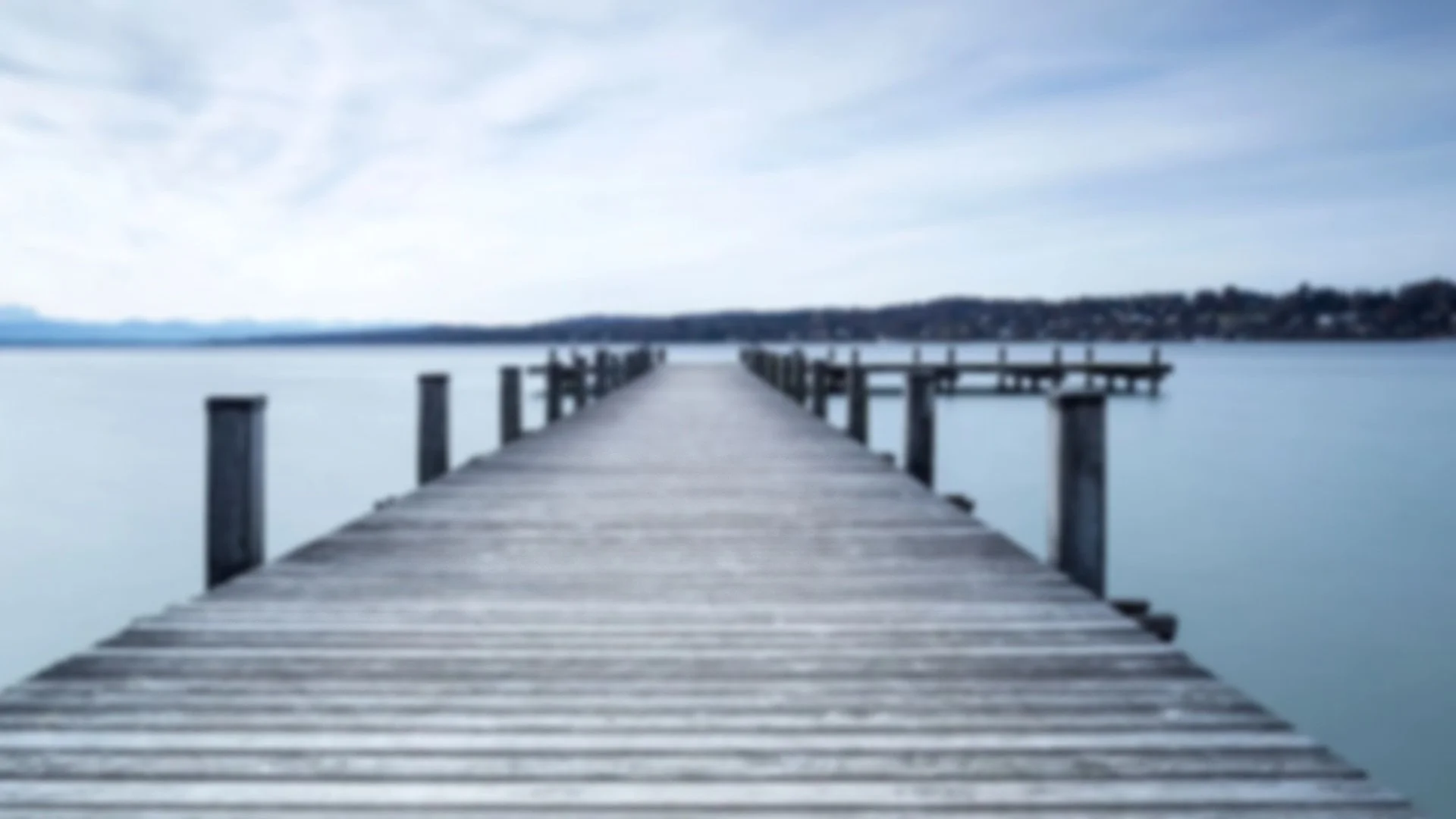 Ein Holzsteg führt über ruhiges Wasser in der Nähe eines Flusses oder Sees, mit Himmel und entferntem Ufer im Hintergrund.