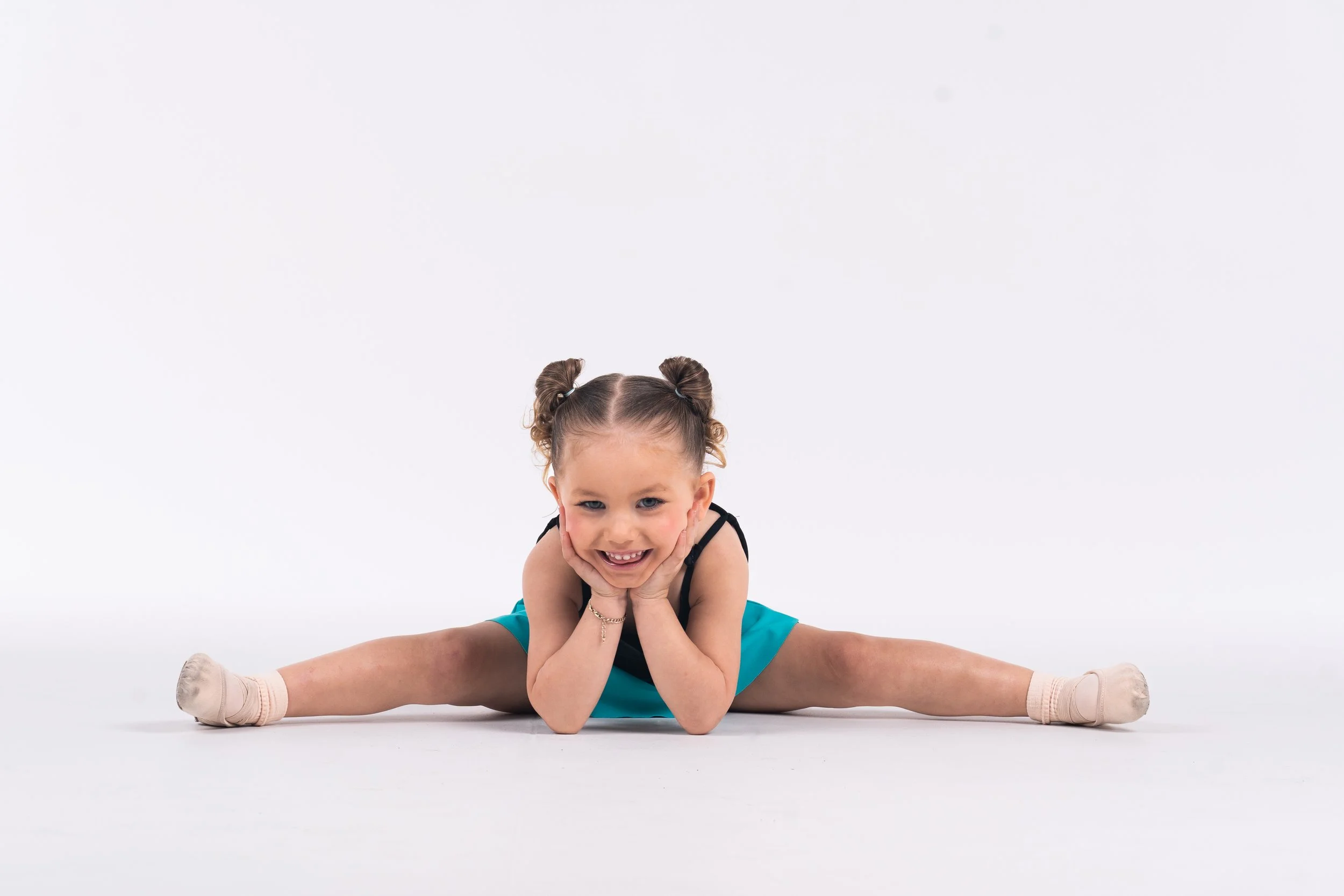 Young girl performing a split, smiling, with knees bent and hands on cheeks, against a plain white background, at the Dance HQ uniform photoshoot.