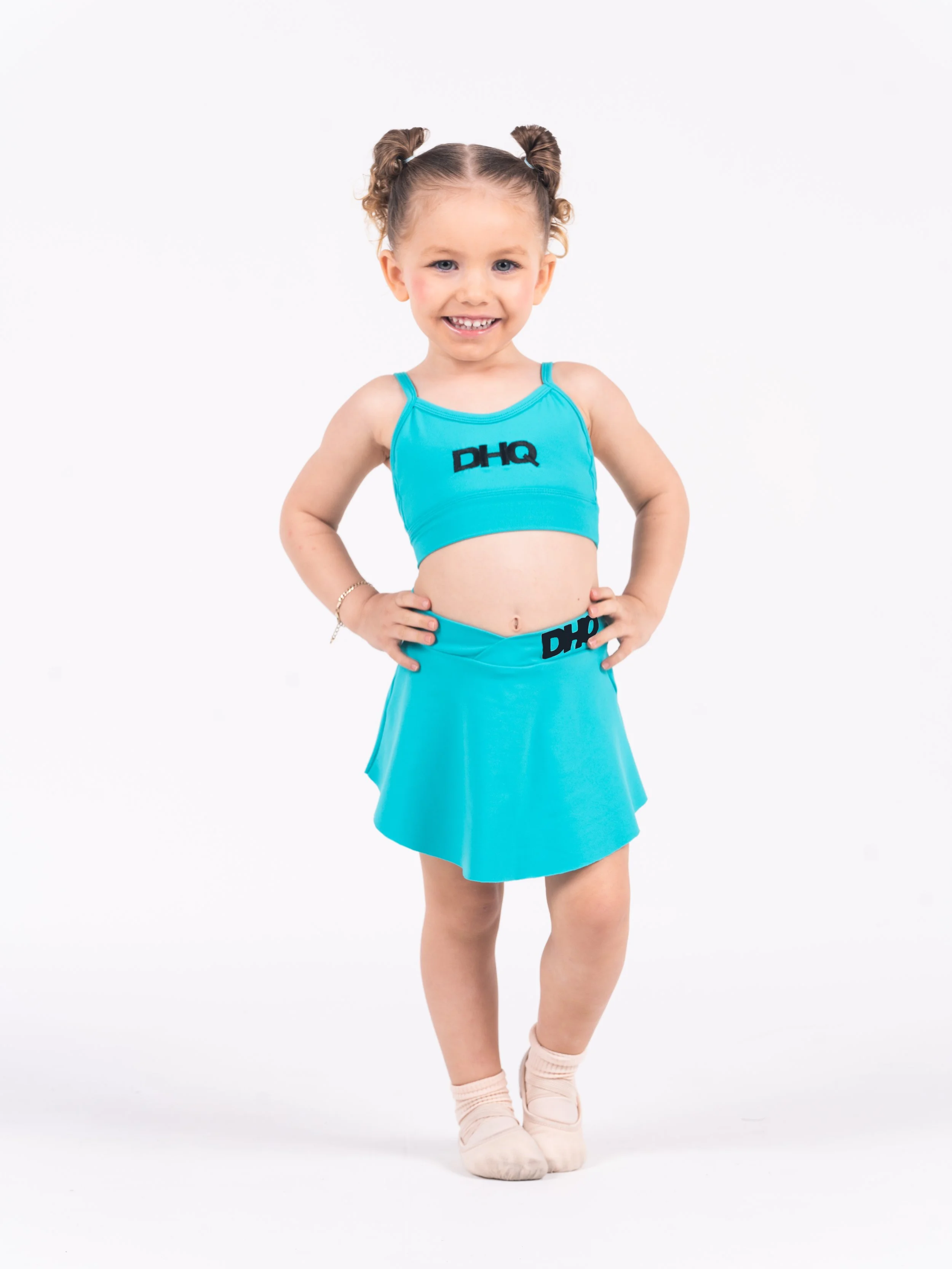 Young girl in a blue sports crop top and matching ballet skirt with the logo 'DHQ' smiling and standing with hands on hips against a plain white background.