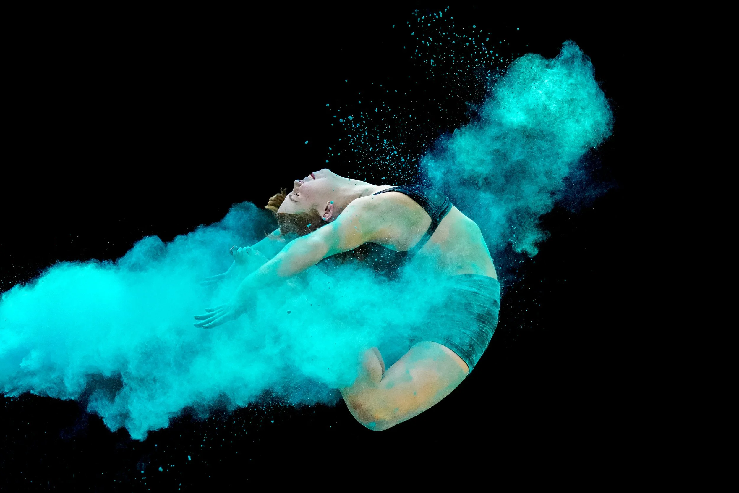 A female Dance Headquarters student dancer is captured mid-air with blue powder exploding around her against a black background.