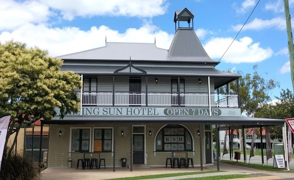 Two-story hotel building with a sign saying 'Living Sun Hotel, Open 7 Days.' The hotel has a porch on the ground floor and a balcony on the second floor, with a large, steep roof and small tower-like structure on top. There are trees and a blue sky with some clouds in the background.