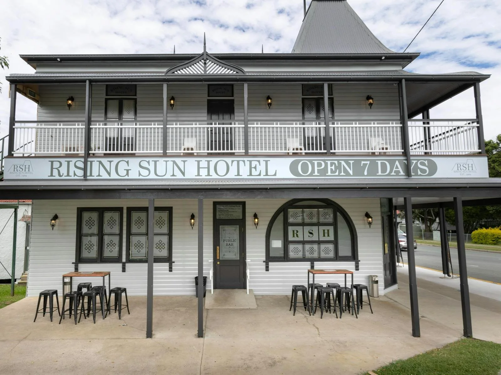 Front of the Rising Sun Hotel, a two-story building with a balcony, black and white color scheme, outdoor seating, and signage indicating it is open 7 days.