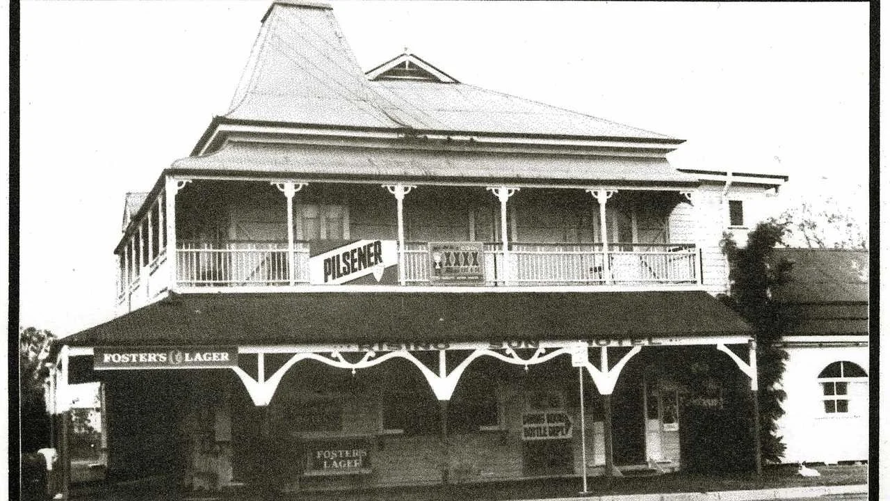 Black and white photo of a two-story house with a large porch and a balcony, advertising Pilsener beer and Foster's Lager, with a sign for XXXX beer and a