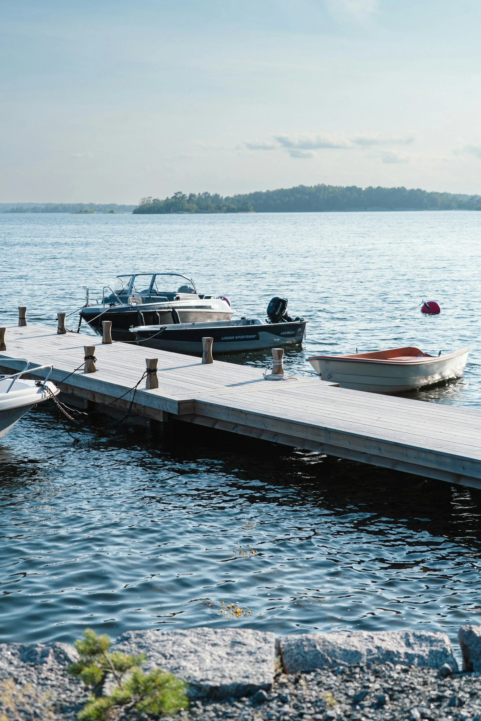 Boats tied to a dock on a lake with a buoy in the water and trees in the background.