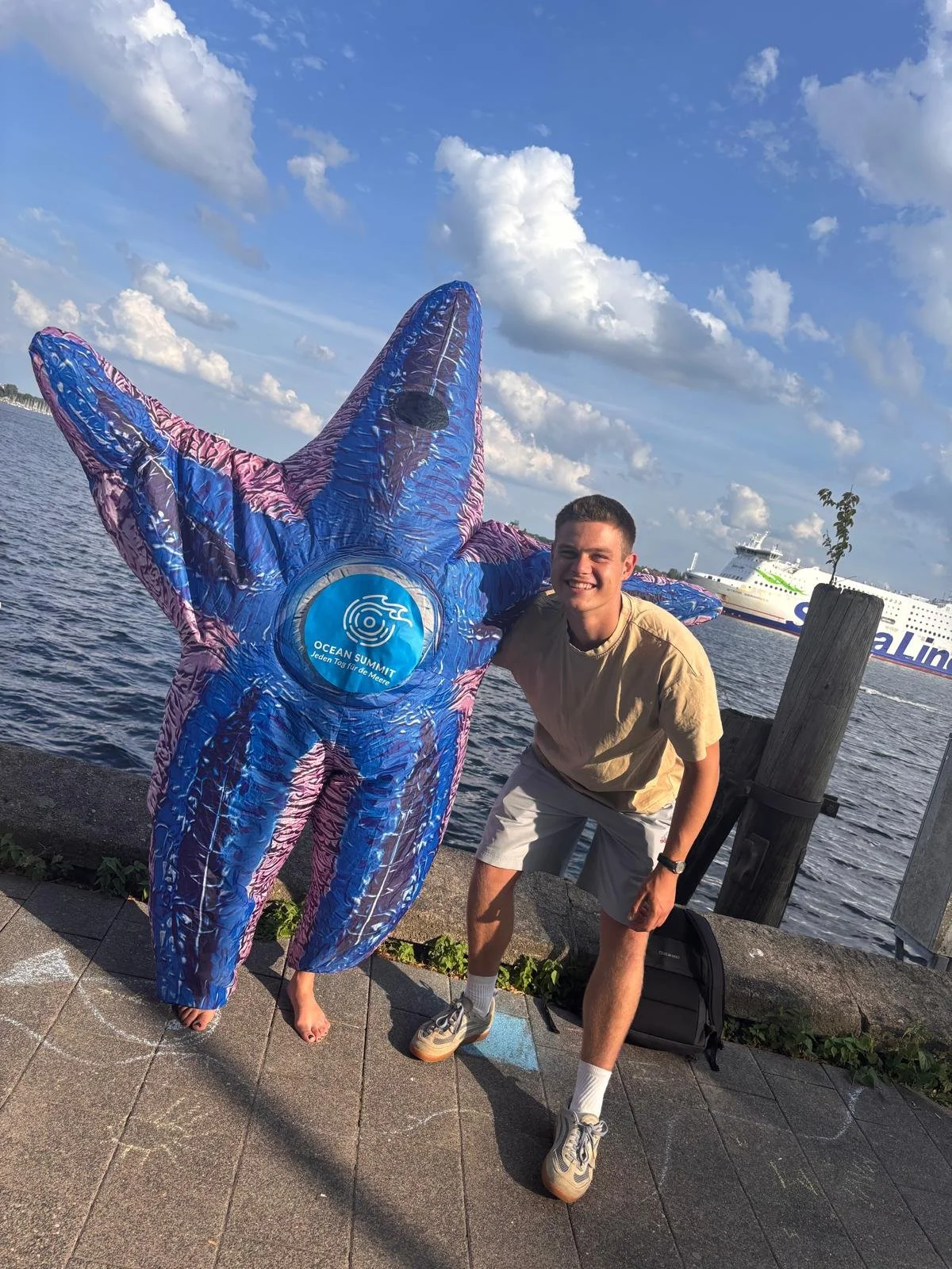 Junge Mann und große, bunte Stern-Skulptur am Wasser mit Schiff im Hintergrund, sonniger Himmel.