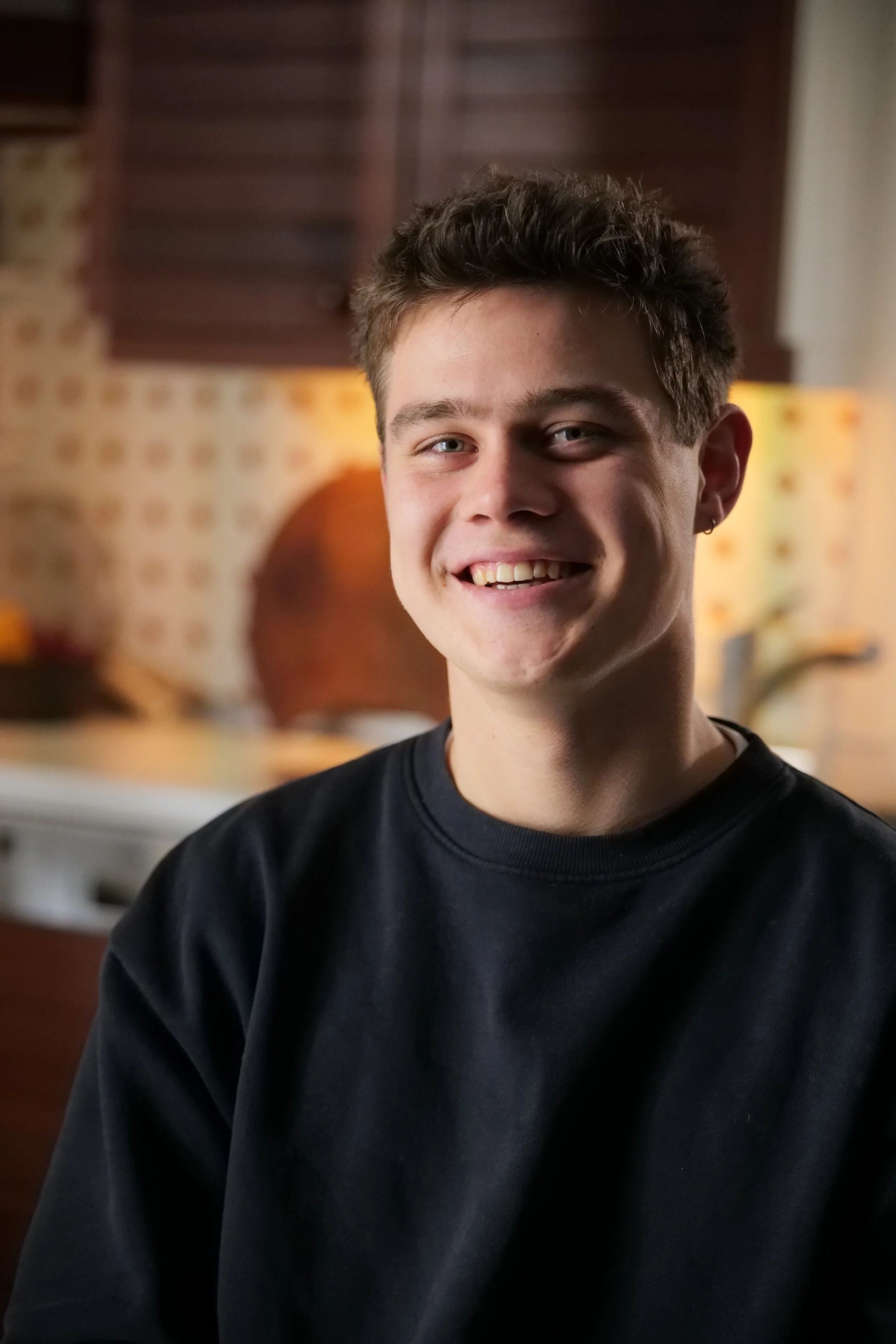 Ein Lächelnder junger Mann in einem schwarzen Sweatshirt vor einer Küchenhintergrund mit rustikalen Holzelementen und buntem Fliesenmuster.