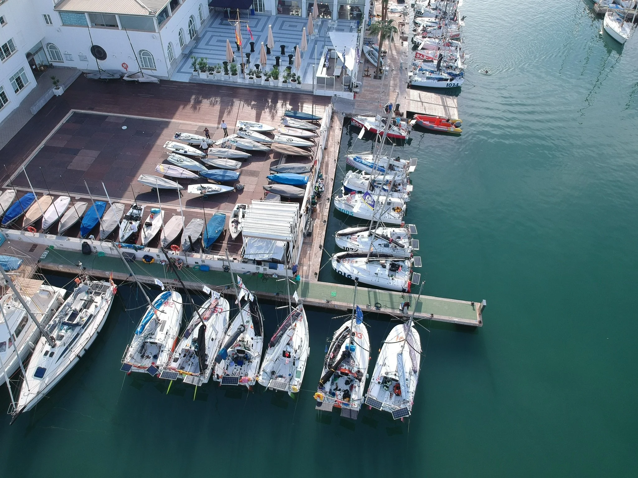 Aufnahme eines Hafens mit mehreren Segelyachten und Booten, die an einem Kai festgemacht sind. Im Hintergrund befindet sich ein Gebäude mit einer Terrasse, Tische und Stühlen, sowie Sonnenschirmen. Das Wasser ist ruhig und grünlich.