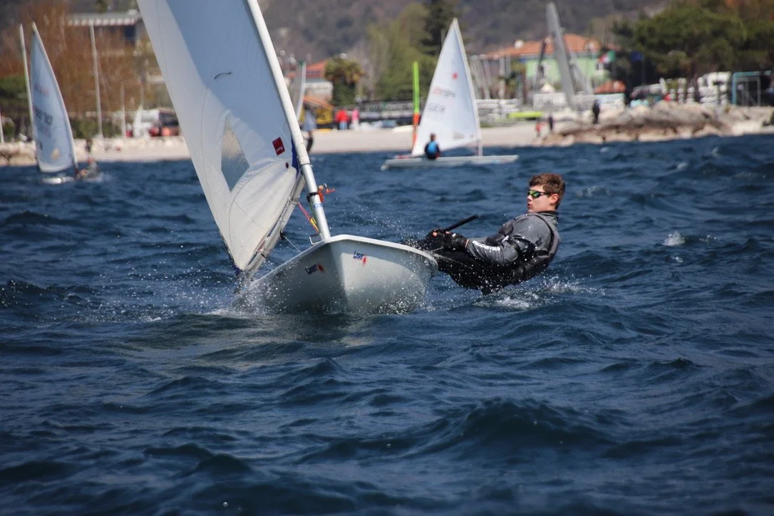 Segler auf einem kleinen Boot beim Windsurfen auf dem Wasser, mit Land und weiteren Seglern im Hintergrund.