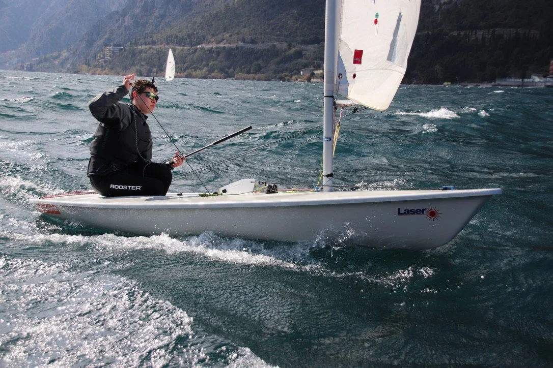 Ein Mann segelt auf einem kleinen weißen Segelboot auf einem See mit Wind und Wellen, im Hintergrund Hügel mit Bäumen und Gebäuden.