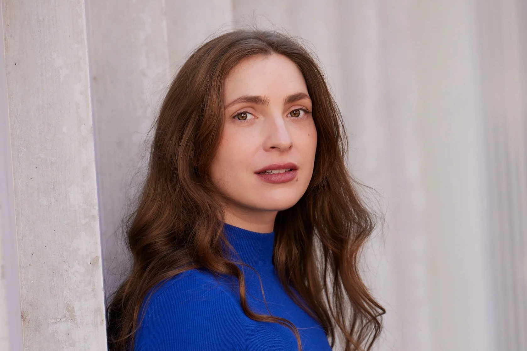 Mela Medina mit langen hell-braunen Haaren und blauen Kleid steht vor einer hellen Wand.