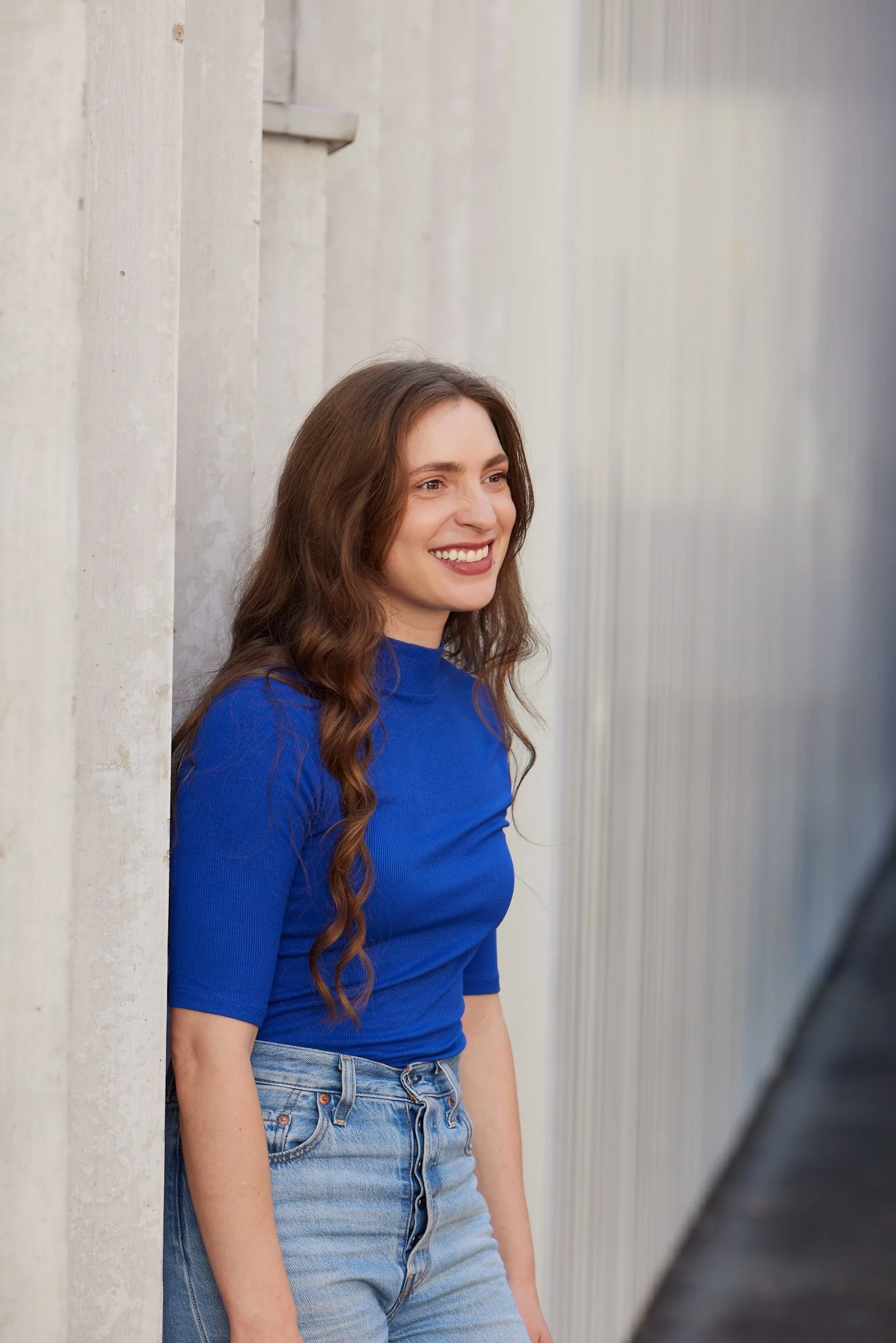 Mela Medina mit hell-braunen, lockigen Haaren, trägt eine blaue langärmlige Oberteil und Jeans, steht an einer Betonwand und lächelt.