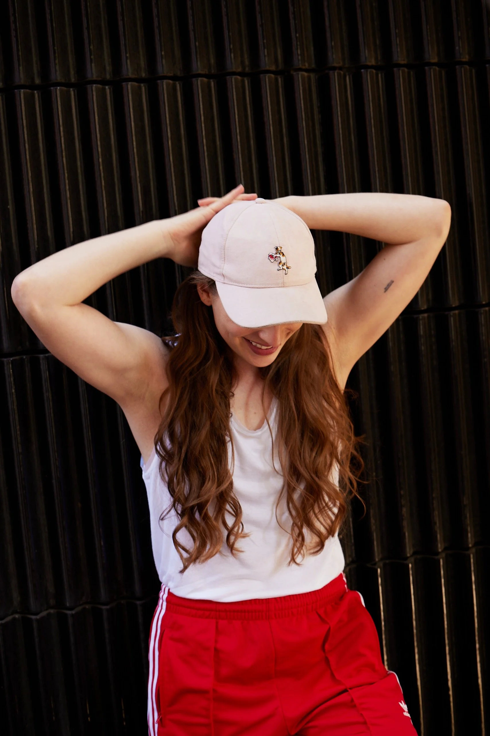 Mela Medina mit langen Haaren, die einen weißen Tanktop und rote Shorts trägt, lächelt und trägt eine weiße Cap mit einem kleinen Katzenmotiv, steht vor einer schwarzen, gestreiften Wand.