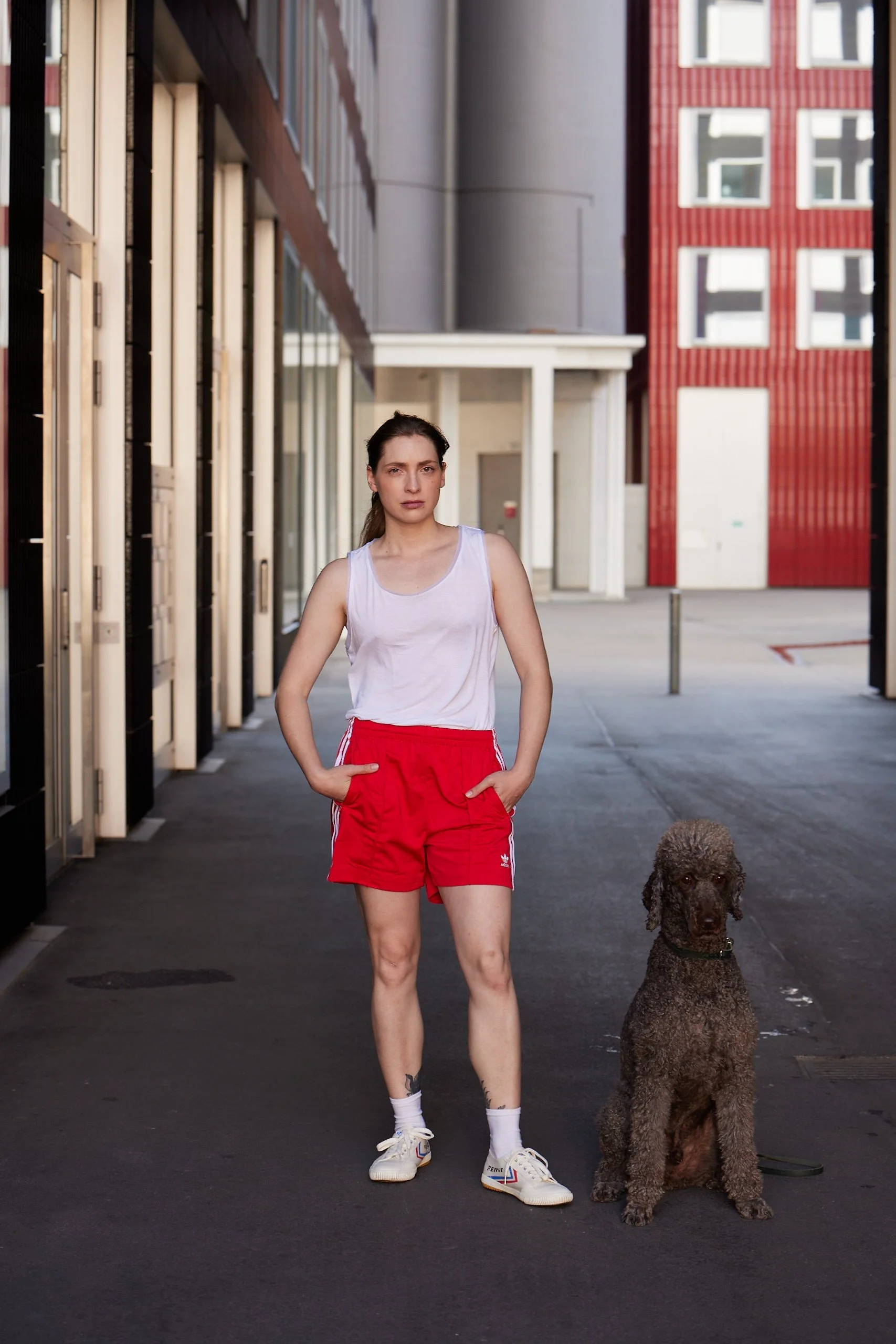 Mela Medina mit hell-braunen Haaren trägt weiße Sportkleidung und roten Shorts, steht auf einer Straße neben einem braunen Pudel, im Hintergrund moderne Gebäude.