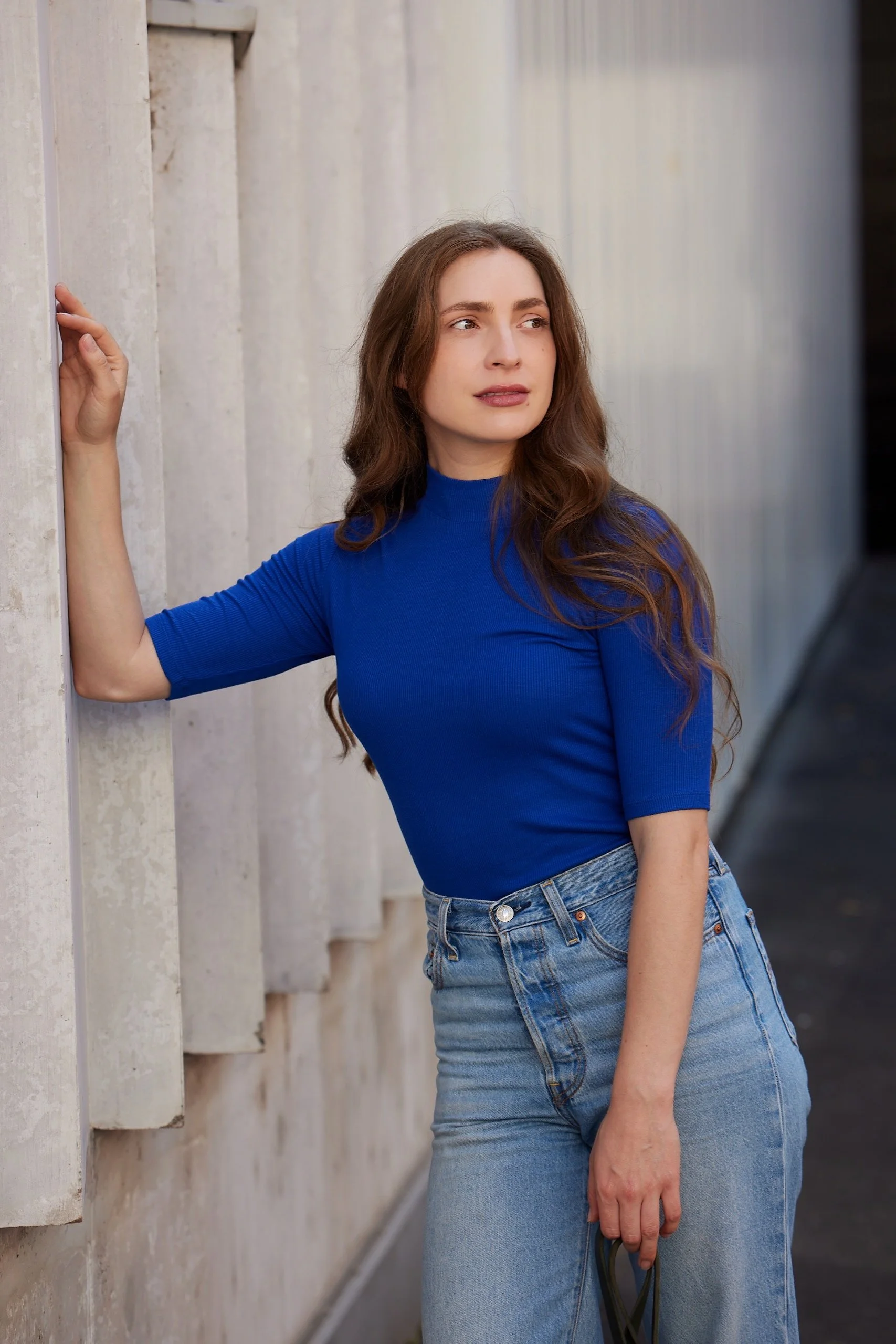 Mela Medina mit hell-braunen Haaren und blauer Oberteil lehnt an einer hellen Wand.