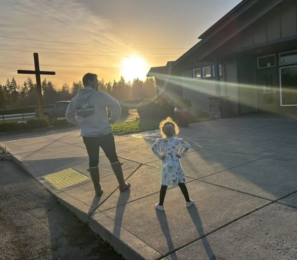 A man and a young girl stand on a concrete driveway, watching the sunset. Sky’s Crew family-owned business in the PNW. There is a wooden cross in the background near a rural landscape with trees and a vintage truck, under a partly cloudy sky.