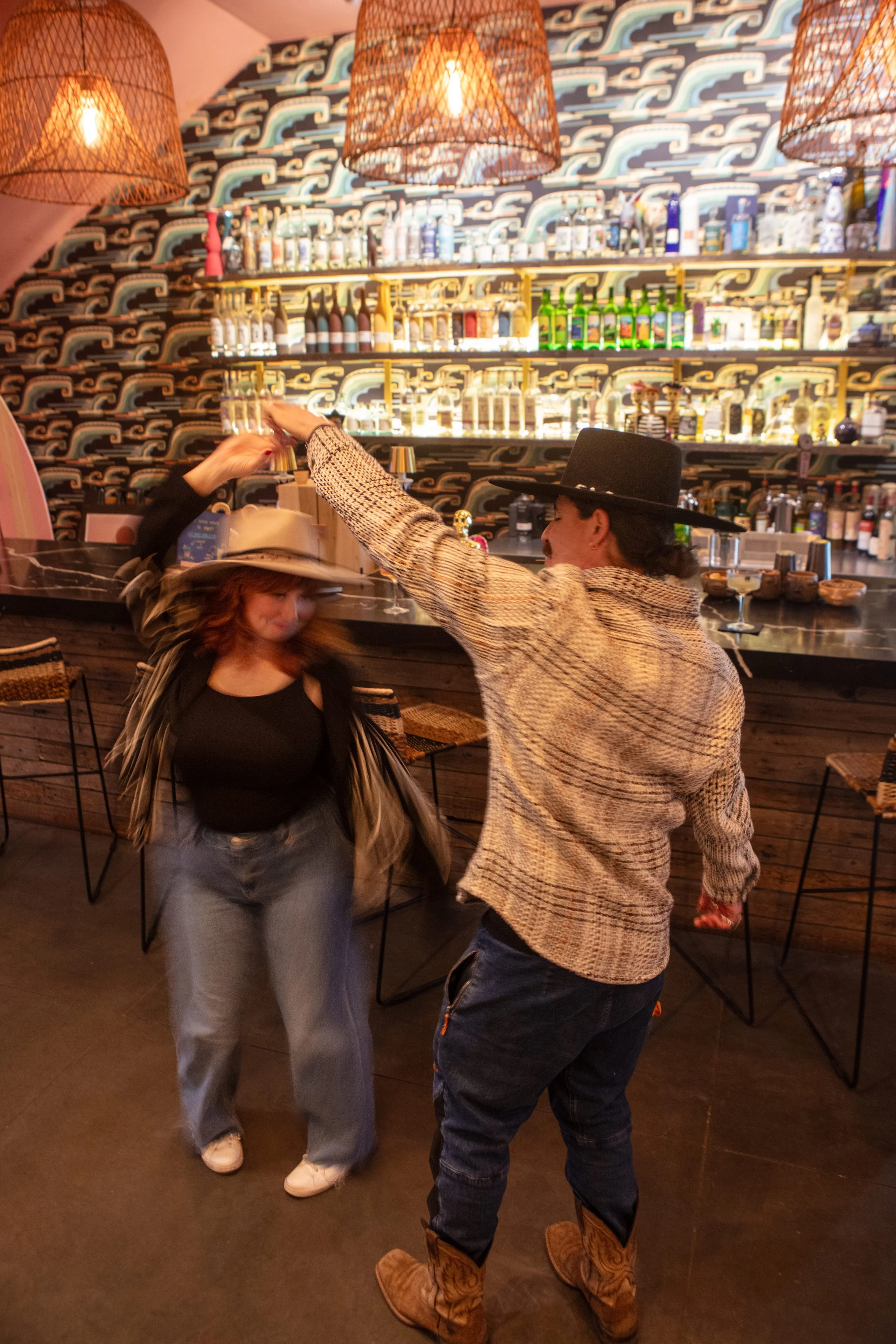 Two people dancing together in a bar or restaurant with a patterned wall and shelves of bottles behind them. One person is a woman with red hair wearing a striped jacket and a beige hat, while the other is a man wearing a wide-brimmed black hat, a checkered shirt, and cowboy boots.