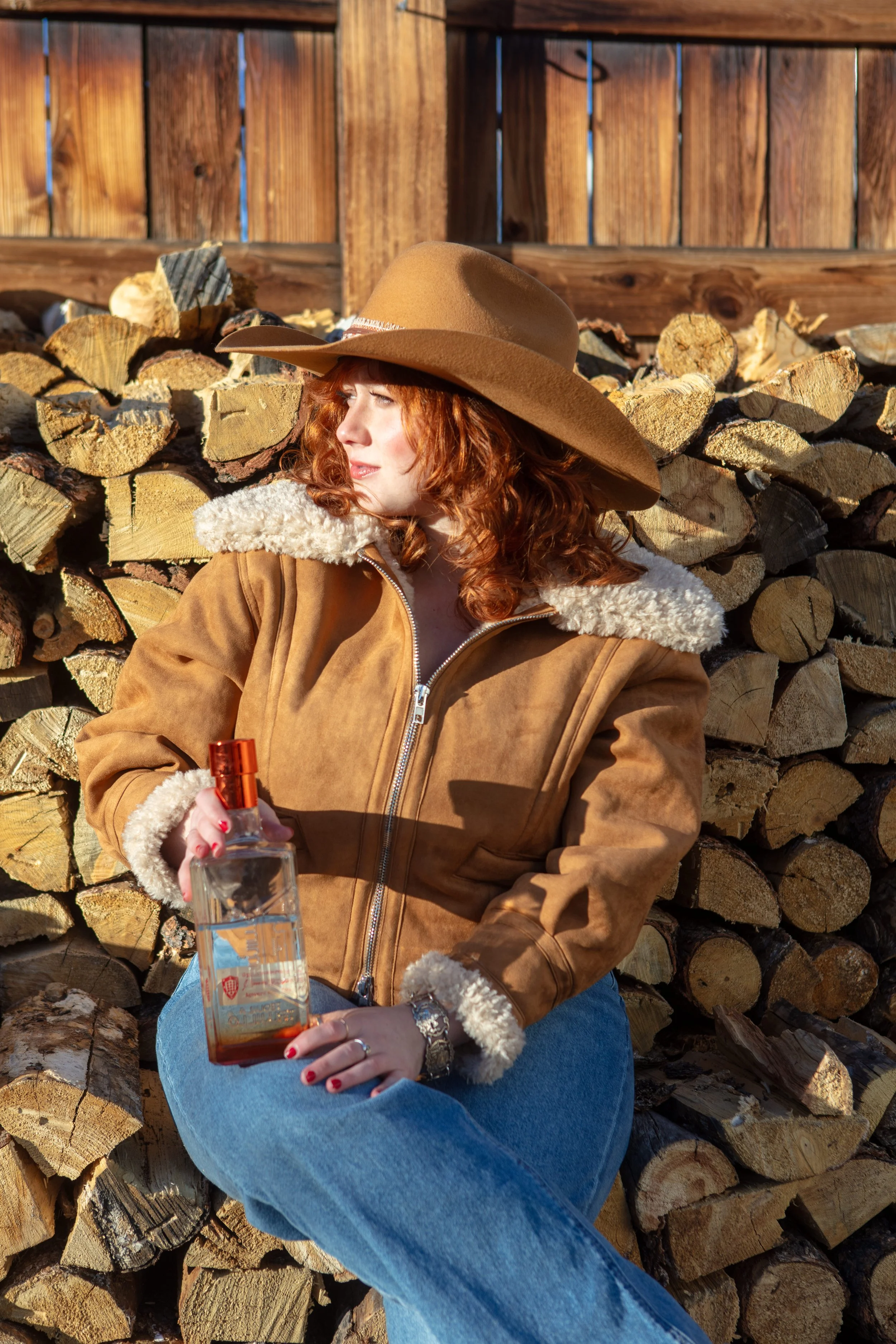 A woman with curly red hair wearing a tan wide-brimmed hat and a brown suede jacket with shearling collar, sitting against a pile of chopped firewood and a wooden fence, holding a bottle of alcohol.