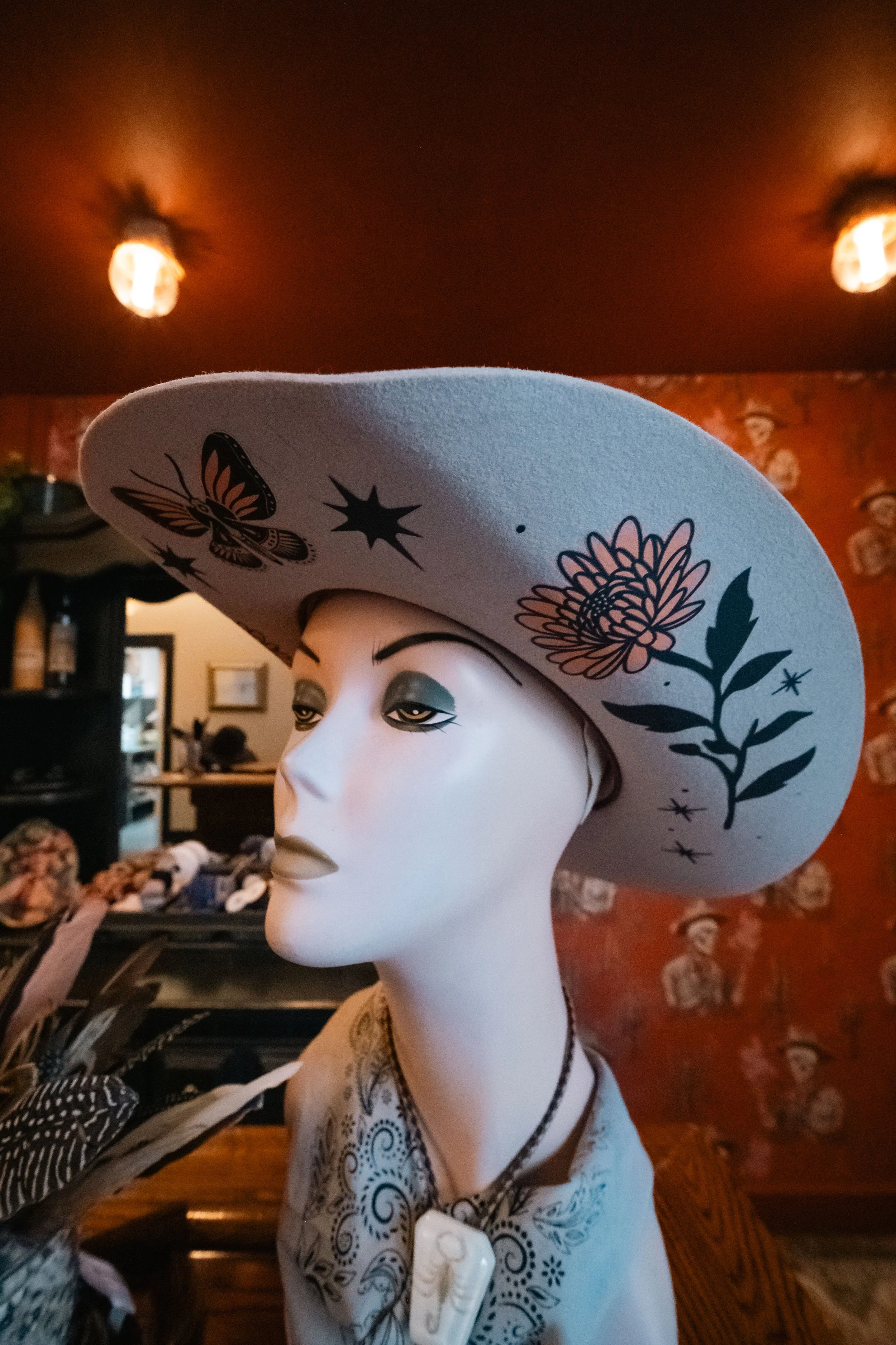 A mannequin wearing a white wide-brimmed hat with black floral and butterfly designs, silver earrings, and a white top with blue intricate patterns inside a store with warm lighting and floral wallpaper.